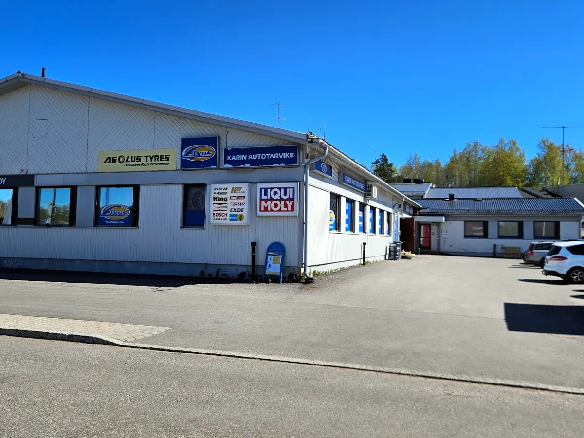 Karin Autotarvikkeen piha — valkoinen liikerakennus, Karin Autotarvike -kyltti, LIQUI MOLY -mainos ja asiakaspysäköinti